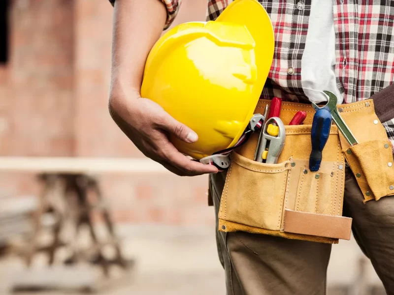 Un entrepreneur général tenant une ceinture à outils et un casque de sécurité sur un chantier de construction.